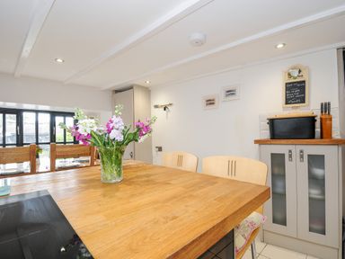 A kitchen with a wooden table with flowers in a glass vase and chairs near a cabinet with a cutting board and knives at Church View Cottage Near Looe