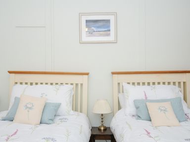 Two single beds with floral bedding and pillows separated by a small table with a lamp and a framed picture on the wall at Church View Cottage Near Looe