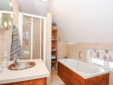A bathroom with a sink vanity a shower stall a bathtub wooden shelving and a window with a roller shade at Church View Cottage Near Looe