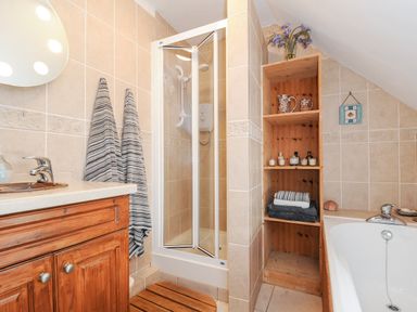 A bathroom with a wooden vanity unit and sink a glass shower enclosure wooden shelves with towels and toiletries and a bathtub with a faucet at Church View Cottage Near Looe