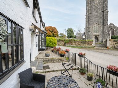 A patio with black metal table and chairs outside a white building near a stone church at Church View Cottage Near Looe