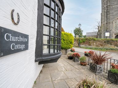 Exterior of Churchview Cottage showing a curved window black window frame potted plants on a stone patio and part of a church building in the background near Looe