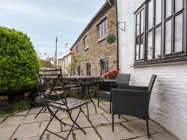 An outdoor patio with metal and wicker chairs a round table and plants near a stone and white building at Church View Cottage Near Looe