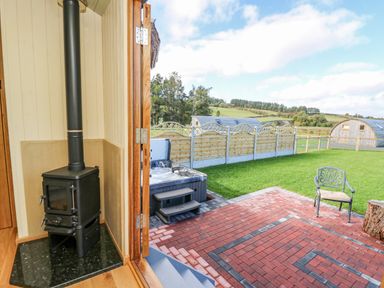 A small wood-burning stove inside a room with an open door leading to a patio with a hot tub a chair and a grassy yard at Poppy in St Harmon near Rhayader