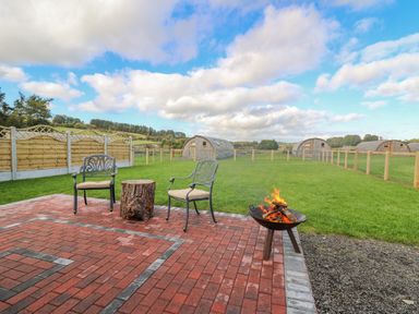 An outdoor patio with two chairs a tree stump table and a fire pit overlooking a grassy field with wooden cabins at Poppy in St Harmon near Rhayader