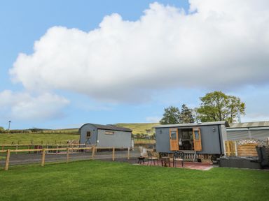 Two gray shepherd huts with wooden doors and windows on a grassy area with fenced fields and hills in the background at Poppy in St Harmon near Rhayader