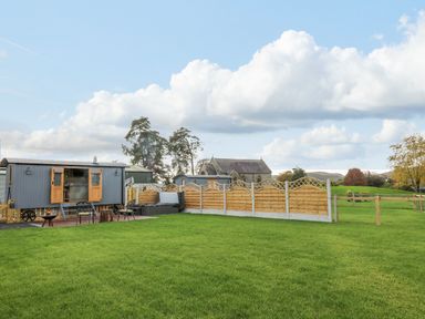 A grassy garden area with a wooden fence a hot tub and two gray caravan-style structures at Poppy in St Harmon near Rhayader