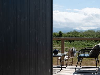 A wooden deck with chairs a table and a teapot overlooking green bushes and mountains at Galwad y Mor in Niwbwrch newborough