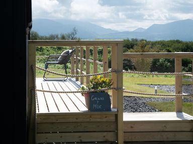 A wooden deck with a chair and a potted plant with a sign near greenery and distant mountains at Galwad y Mor in Niwbwrch newborough