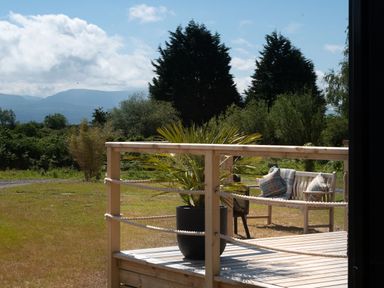 A wooden deck with a potted plant and a bench with cushions and a blanket in a garden with trees and mountains in the background at Galwad y Mor in Niwbwrch newborough