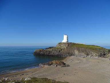 A small white tower on a rocky hill by the sea with a sandy beach in the foreground at Galwad y Mor in Niwbwrch newborough