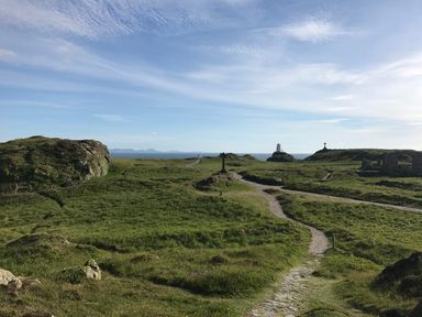 A coastal landscape with grassy terrain and walking paths leading to a lighthouse cross and ruins at Galwad y Mor in Niwbwrch newborough