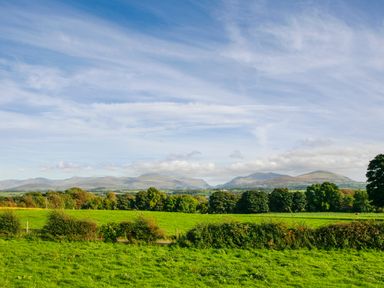 A rural landscape with green fields trees and hills under a blue sky at Galwad y Mor in Niwbwrch newborough