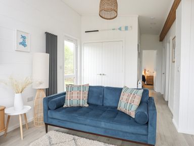 A living room with a blue sofa two patterned cushions a white side table with a vase and dried plants a tall lamp and a paddle mounted on the wall at Galwad y Mor in Niwbwrch newborough