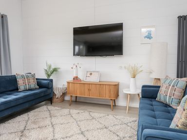 A living room with two blue sofas a wooden TV stand a wall-mounted TV and a white side table with a lamp at Galwad y Mor in Niwbwrch newborough