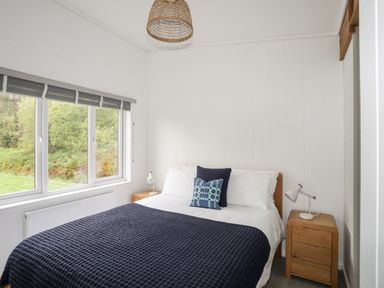 A bedroom with a bed featuring blue blankets and two cushions beside two wooden nightstands with lamps at Galwad y Mor in Niwbwrch newborough