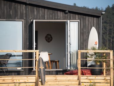 A wooden deck in front of a dark house with open double doors showing a table and chairs inside a surfboard and skateboard on the deck with a potted plant at Galwad y Mor in Niwbwrch newborough