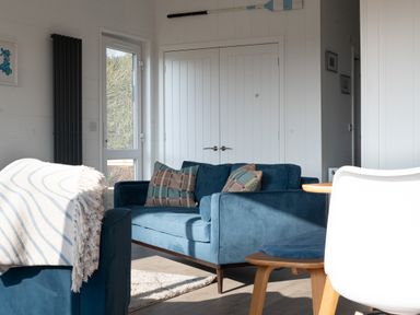 A living room with blue sofas and patterned cushions near white double doors at Galwad y Mor in Niwbwrch newborough