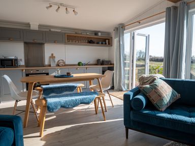 A kitchen dining area with wooden table chairs and bench blue cushions and sofa near glass doors at Galwad y Mor in Niwbwrch newborough