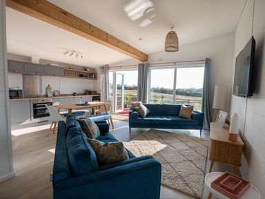 A living room with two blue sofas a rug and a wooden cabinet next to a kitchen with a dining table and chairs at Galwad y Mor in Niwbwrch newborough