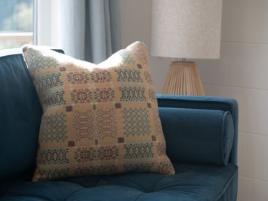 A patterned cushion on a blue sofa with a lamp and curtains in the background at Galwad y Mor in Niwbwrch newborough
