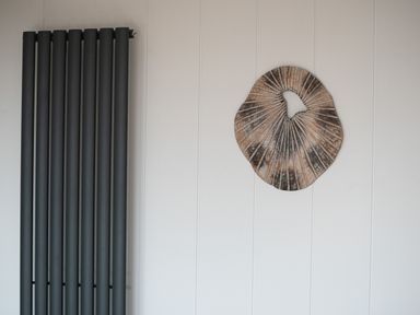 A wall with a vertical black radiator and a round wooden decorative piece at Galwad y Mor in Niwbwrch newborough