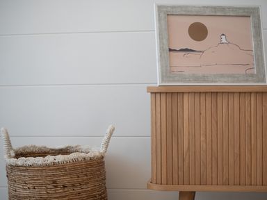 A wicker basket next to a wooden cabinet with a framed illustration on top at Galwad y Mor in Niwbwrch newborough