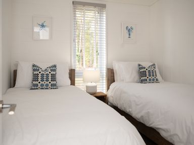 A bedroom with two single beds a window with blinds and a lamp on a wooden side table between the beds at Galwad y Mor in Niwbwrch newborough