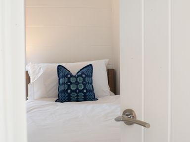A bed with white sheets and pillows with a blue patterned cushion in a bedroom seen through a partially open white door at Galwad y Mor in Niwbwrch newborough
