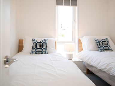 A bedroom with two single beds white bedding and blue patterned pillows and a bedside table with a lamp at Galwad y Mor in Niwbwrch newborough