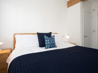 A bedroom with a wooden bed frame white bedding navy blue blanket two blue cushions bedside tables with lamps at Galwad y Mor in Niwbwrch (newborough)