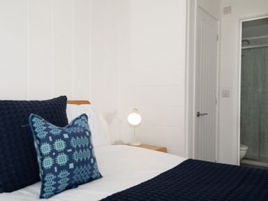 A bedroom with a bed with blue and patterned cushions a bedside table with a lamp and an open door showing a bathroom at Galwad y Mor in Niwbwrch newborough