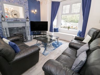 A living room with black leather sofas a glass coffee table a fireplace and a large window with blue curtains at Gwyder in Penmaenmawr