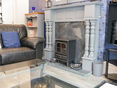 A living room with a leather sofa and a blue cushion a wooden shelf with books a black stove in a fireplace and a glass coffee table at Gwyder in Penmaenmawr