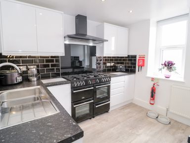 A kitchen with black countertops black tiled backsplash white cabinets a black oven a stainless steel sink a toaster a fire extinguisher and flower vase on the window at Gwyder in Penmaenmawr