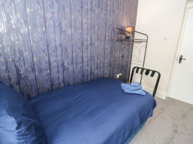 A bedroom with a single bed covered in blue bedding and folded towels on top near a white door with a metallic rack and lamp against a patterned accent wall at Gwyder in Penmaenmawr
