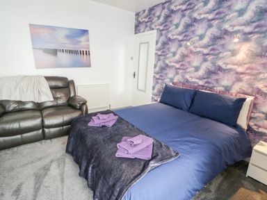 A bedroom with a double bed blue bedding purple towels a dark sofa and a wall with purple cloud pattern at Gwyder in Penmaenmawr