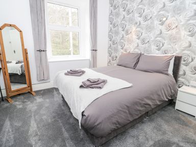 A bedroom with a double bed grey bedding floral wallpaper a full-length mirror and grey curtains at Gwyder in Penmaenmawr