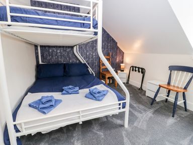 A bedroom with a bunk bed with blue bedding towels on the bed two chairs two white bedside tables a lamp and a luggage rack at Gwyder in Penmaenmawr