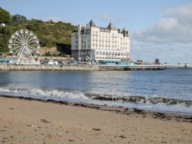 A sandy beach with waves and a pier leading to a large building with towers and a ferris wheel near the water at Gwyder in Penmaenmawr