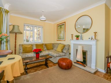 A living room with a patterned sofa and yellow cushions a wooden coffee table with a red flower pot a fireplace with a large round mirror above it and a woven rug on the floor at See The Sea in Hunstanton