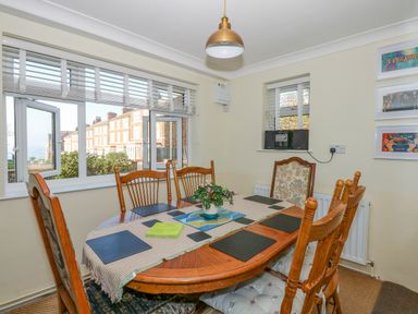 A dining room with a wooden table and six chairs next to windows overlooking houses and greenery at See The Sea in Hunstanton