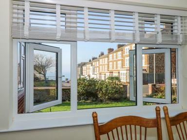 A window with white blinds overlooking a street with brick houses and greenery at See The Sea in Hunstanton