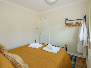 A bedroom with a bed covered with a mustard yellow quilt and two rolled white towels on it a white door and a wall-mounted rack with wooden hangers and white robes at See The Sea in Hunstanton