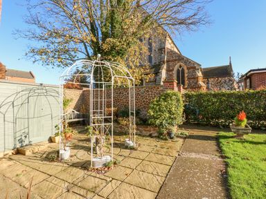An outdoor garden area with a white metal gazebo and stone paving near a brick and stone church at See The Sea in Hunstanton