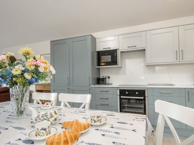 A kitchen with blue and white cabinets a microwave an oven a table set with croissants tea cups and a vase of flowers at Lambley View in Bude