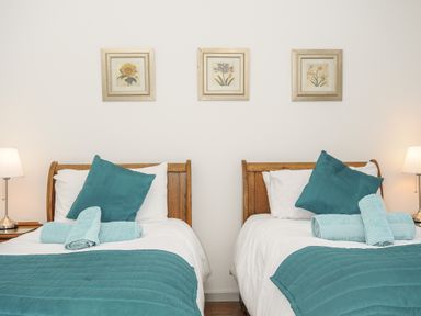 A bedroom with two single wooden beds each with teal bedding and pillows and rolled towels on the beds with lamps on each side and three floral pictures on the wall at Lambley View in Bude