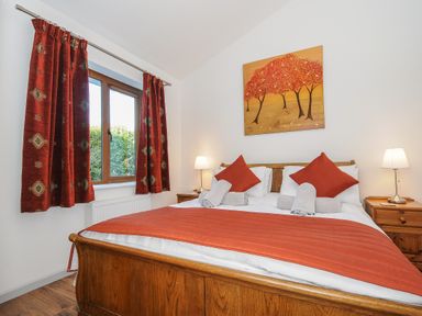 A bedroom with wooden bed frame orange bedding and pillows bedside tables with lamps and painting of trees on wall at Lambley View in Bude