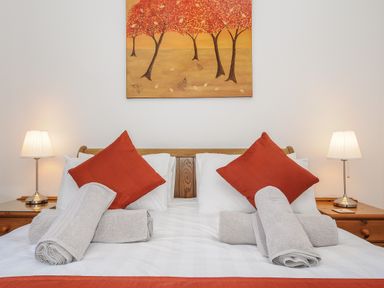 A bedroom with a double bed with red pillows and rolled towels on white sheets with two bedside tables and lamps and a painting of trees above the bed at Lambley View in Bude