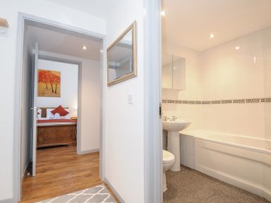 A hallway with a mirror on the wall leading to a bedroom with a bed and artwork and a bathroom with a sink and bathtub at Lambley View in Bude